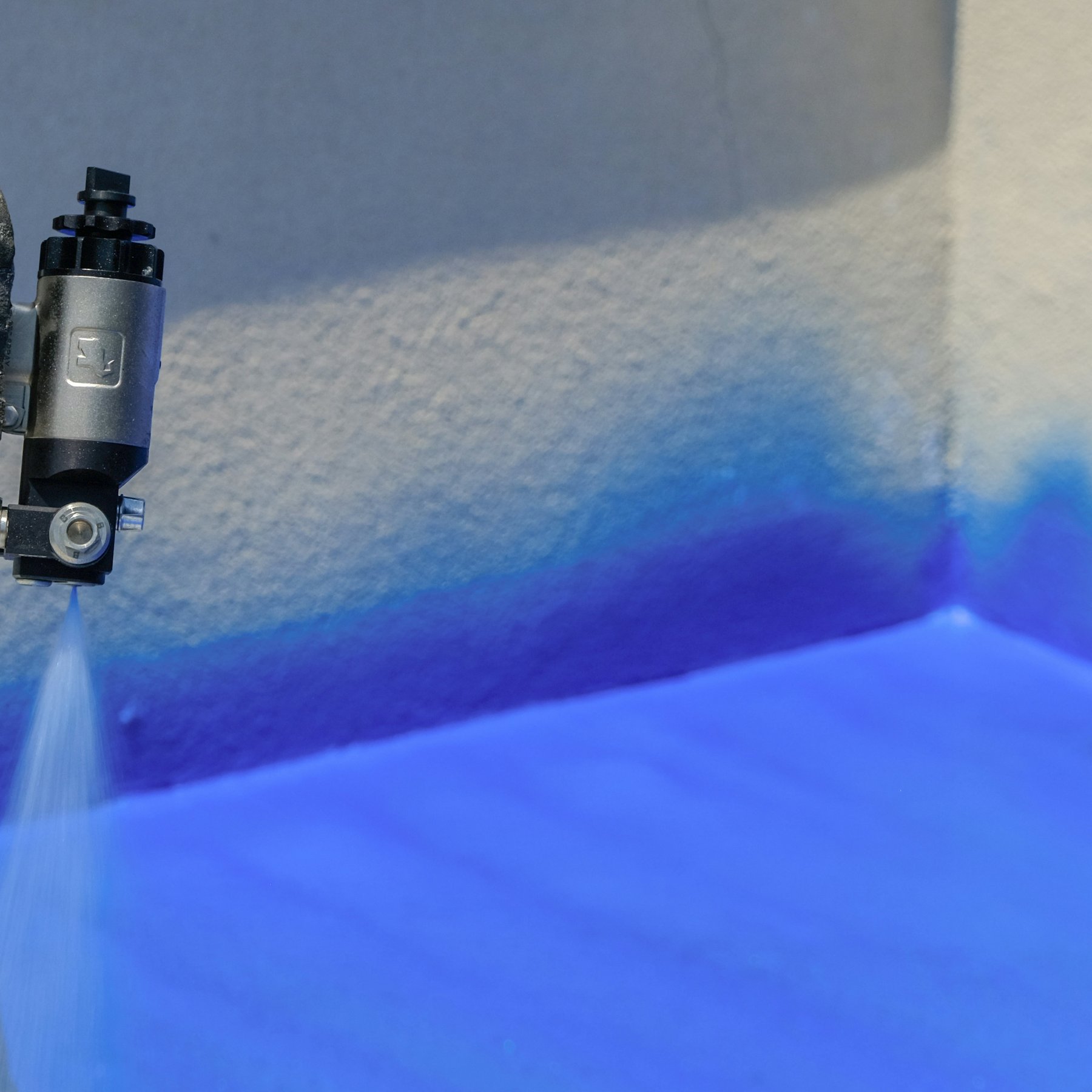 Spray gun applying a blue liquid coating to a wall and floor junction